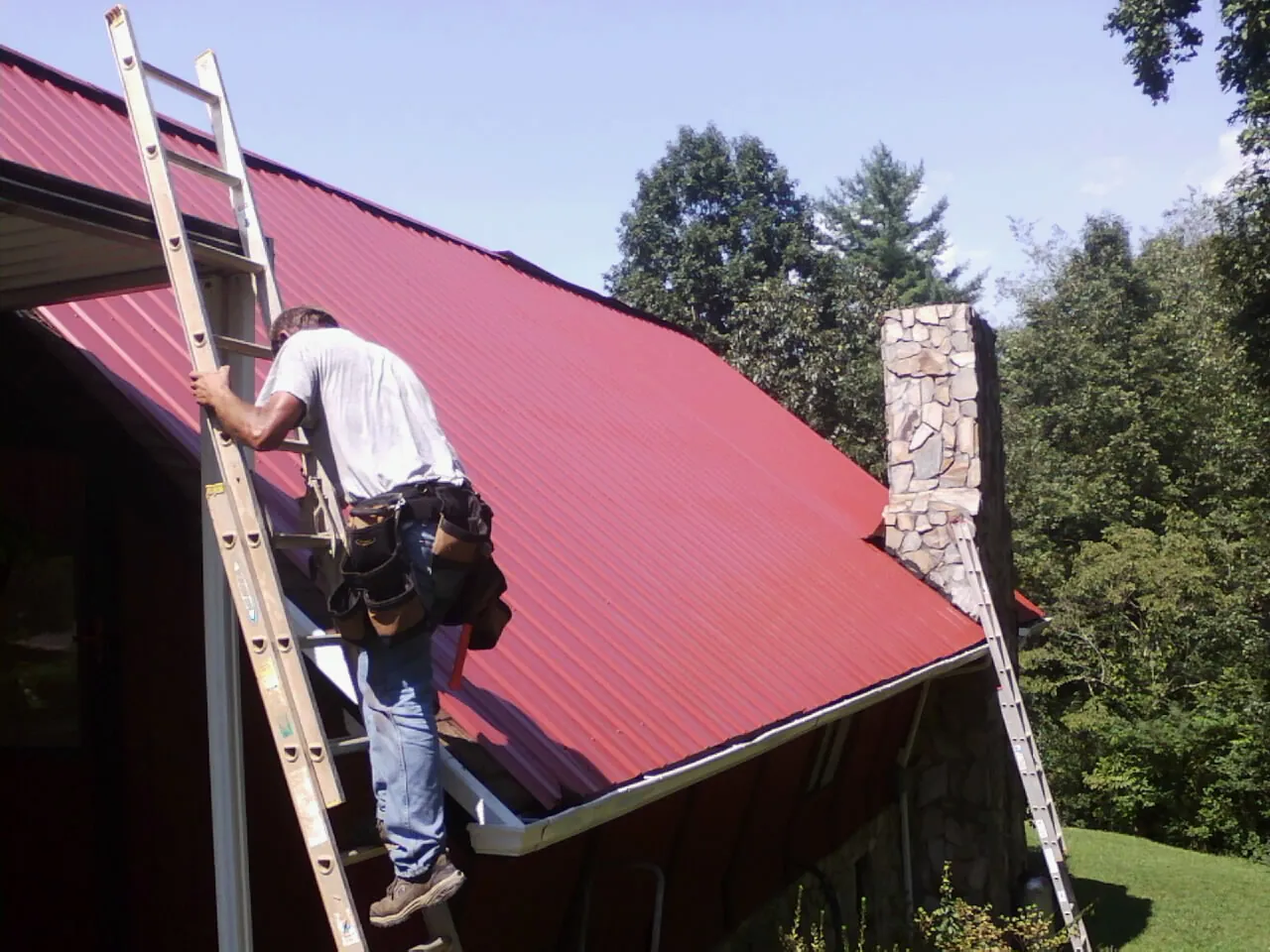 Roofer on a ladder beside a red metal roof and stone chimney — Soffit & Fascia Repair by Roofing Brothers in Wise